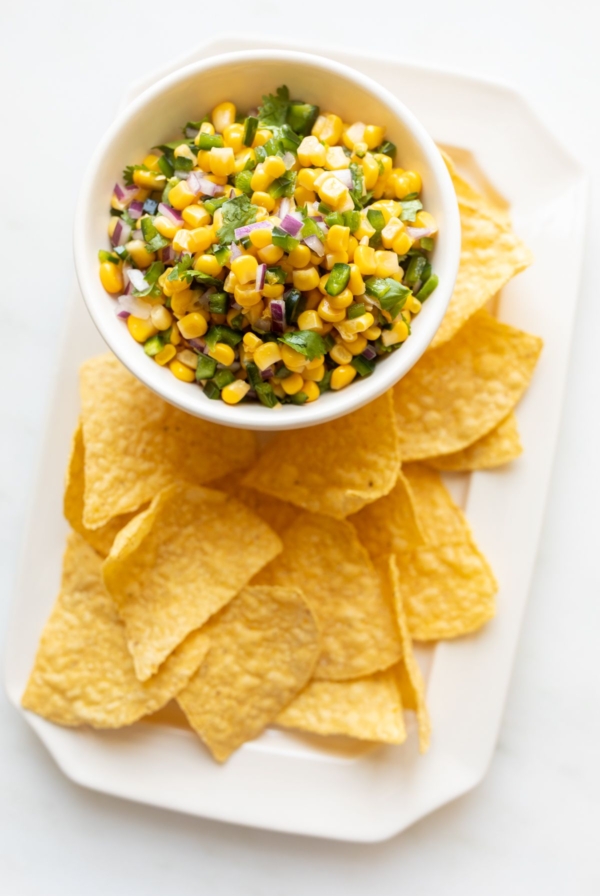 A chipotle corn salsa recipe in a white bowl, placed on a white platter full of corn tortilla chips