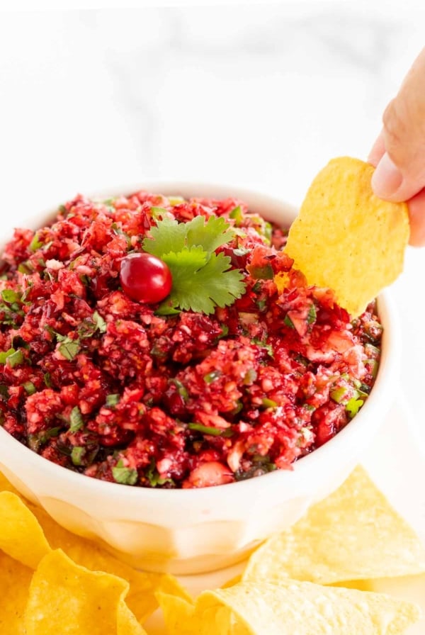 A hand dipping a tortilla chip into a bowl of fresh Christmas cranberry salsa.