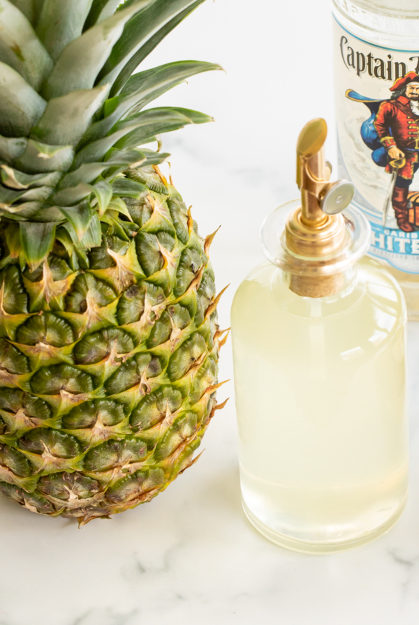 Bottle of pineapple simple syrup set next to fresh pineapple and bottle of white rum